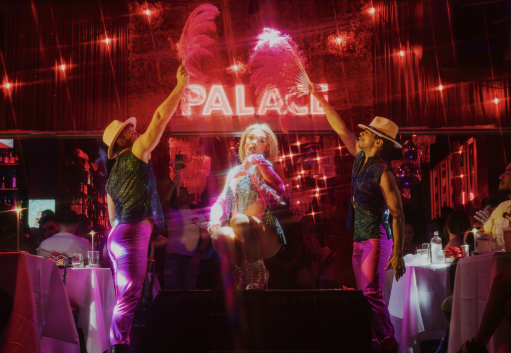 Drag queen performing with two dancers at Palace South Beach in Miami Beach, beneath the iconic Palace neon sign.